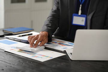 A focused businessman sits at his office desk, analyzing a paper chart while working on his laptop. He examines financial and marketing reports, concentrating on data for strategic decision-making.