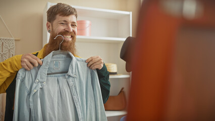 Redhead bearded man smiles holding a shirt in a tailor shop, reflecting a casual handsome adult vibe indoors.