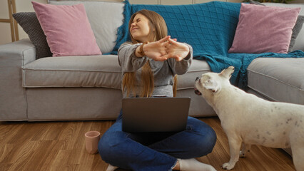 A young, blonde woman stretches while sitting on the floor with her laptop and a coffee cup beside her, a cute bulldog standing nearby in a cozy living room.