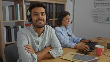 Man and woman working together in an office wearing headsets indicating they may be involved in customer service or support roles, with office supplies and books on shelves in the background