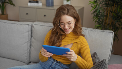 Poster - A young, attractive blonde woman sits comfortably in her living room, smiling while using a blue tablet on a gray couch, surrounded by indoor plants and home decor.