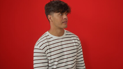 A handsome young hispanic man in a striped shirt is standing against a vibrant red background, looking to the side with a thoughtful expression.