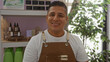 © Krakenimages.com - Young hispanic man indoors in a bakery wearing a brown apron smiling at the camera surrounded by plants and shelves with products