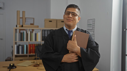 Hispanic man wearing judge's robe holding a book in an office environment, suggesting a legal profession, with bookshelves and office supplies visible in the background.