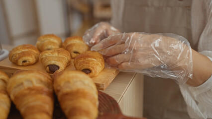 Woman handling freshly baked croissants with plastic gloves in a bakery shop interior with wooden trays displayed.