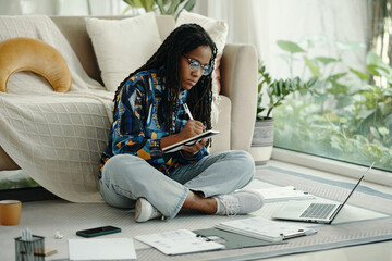 African American woman writing in notebook while sitting on floor in modern living room surrounded by laptop and paperwork, deeply focused on task at hand