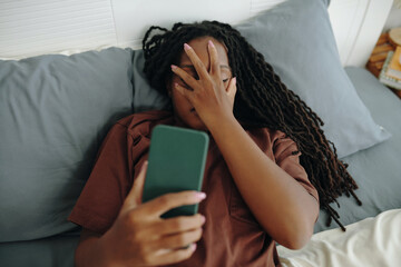 Woman reclining on comfortable bed, holding smartphone in one hand, resting in cozy bedroom filled with soft pillows, displaying relaxed and peaceful demeanor