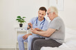 © Studio Romantic - Senior patient learning to use a health care app on a mobile phone. Chubby overweight old man and a friendly young doctor in a uniform with a stethoscope sitting on a bed using a modern phone together