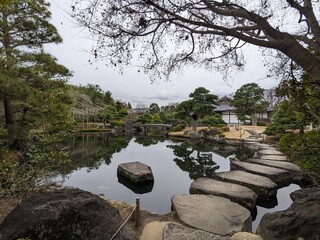 Himeji, Japan - 12.19.2023: A stone pathway over a calm pond leading to a traditional Japanese garden in Koko-en surrounded by manicured trees under a cloudy sky