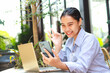 © M Alfan Setyawan - excited asian woman sitting in outdoor cafe with laptop on table holding smartphone showing ok signs say excellent work