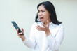 © M Alfan Setyawan - disgruntled furious asian woman looking to smartphone with screaming dissatisfied and raising palm, speaking and questioning gesture, wearing white formal suit standing over isolated background
