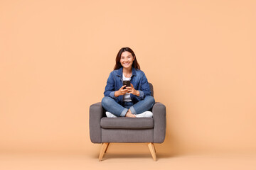 Smiling woman with smartphone sitting in armchair on beige background