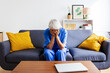© Xavier Lorenzo - Mature female nurse in blue scrubs covering face with hands feeling overwhelmed sitting on sofa after hard work day in hospital. Sad female healthcare worker resting at home. Health care lifestyle
