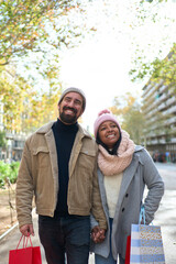 Vertical photo. Beautiful young couple interracial in love enjoying winter holiday season in the city streets, taking a walk, hugging and holding gift bags while celebrating Christmas outdoors