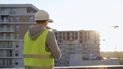 Man constructive engineer with white hard hat and safety vest is using a tablet computer while inspecting a construction site at sunrise in early morning