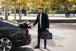 © Aleksandr - Businessman takes a bag and closes trunk of his luxury car and go to airport. Stylish bearded man in coat on auto parking
