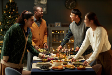 Wall Mural - Side view of cheerful Black man serving dishes on dining table with variety of festive food, while talking with Asian male friend at gathering for Christmas dinner