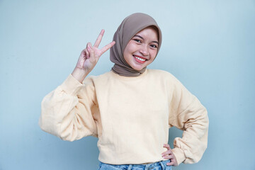 Beautiful smiling Asian woman in a cream sweater looking at camera and showing peace sign with fingers isolated over gray background.