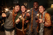 © Seventyfour - Medium full shot of cheerful multiethnic friends holding wine glasses and bright sparklers looking at camera while celebrating Christmas together in living room at home