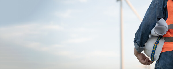 A person wearing a hard hat and orange vest is standing in front of a wind turbine. Concept of hard work and dedication, as the person is likely a worker in the field of renewable energy