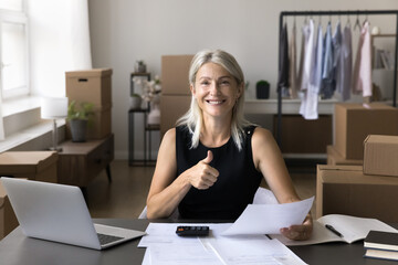 Headshot portrait happy smiling elderly female freelancer looking at camera with thumb up gesture holding finance report. Online store owner satisfied with high income level getting financial profit