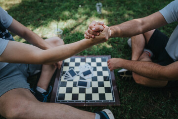 Two friends engage in arm wrestling outdoors, sitting on grass beside a chess board, enjoying a fun and competitive moment.