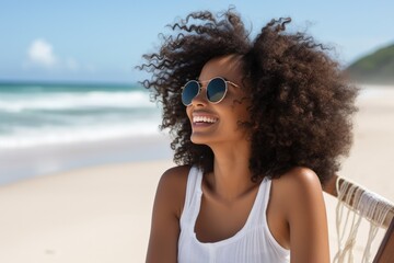 Woman with curly hair is sitting on a beach chair, wearing sunglasses and smiling. Concept of relaxation and enjoyment of the beach environment