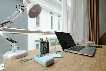 Modern workspace featuring open laptop, desk lamp, and organized stationery on a wooden desk next to a large window with urban view suggesting productivity and focus