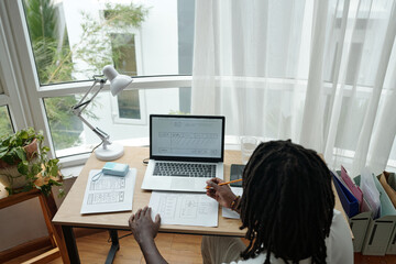 Wall Mural - multiracial woman sitting at desk examining architectural plans on laptop screen. bright workspace n