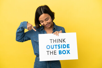 Young mixed race woman isolated on yellow background holding a placard with text Think Outside The Box and  pointing it