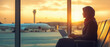 © Lubo Ivanko - Middle eastern woman waiting at airport hall, working on a laptop, airplane visible behind window during evening sunset light. Generative AI