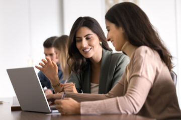 Two cheerful young Hispanic business coworkers using laptop at work table, talking and laughing, discussing ideas for marketing strategy, Internet startup, studying online together