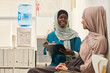 © pressmaster - Smiling Muslim healthcare professional wearing scrubs and a headscarf engaging with a patient seated in waiting room near water cooler and shelves with files
