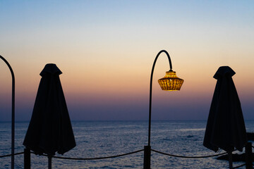 A serene seaside sunrise with a warm, illuminated lantern hanging between two closed umbrellas. The sky fades from soft orange to deep blue, reflecting the peaceful ambiance of the coastal setting.