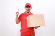 © ArvStd - Happy asian delivery courier in red t-shirt and cap carrying a box package while showing one finger. isolated over white background