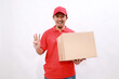 © ArvStd - Happy asian delivery courier in red t-shirt and cap carrying a box package while showing three fingers. isolated over white background
