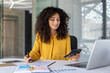© Liubomir - Latin American businesswoman accountant at office desk with calculator, pen, and laptop, demonstrating multitasking skills in financial work. Professional woman exemplifies confidence and efficiency.