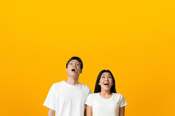 Excited asian man and woman looking upwards at copy space, yellow studio background