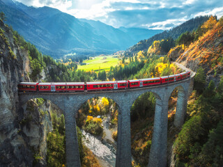 Wall Mural - aerial view of modern red train on landwasser viaduct in alpine mountains, orange forest at sunset i