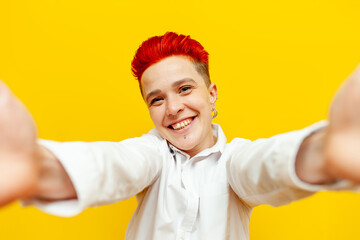 young short-haired red-haired girl hugs and raises her hands and shows welcome gesture on yellow isolated background, friendly hipster woman with piercing holds camera and takes selfie