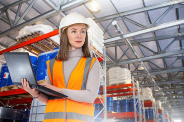 Poster - Woman storekeeper. Girl works in industrial warehouse. Storekeeper with laptop in industrial building. Warehouse racks with barrels behind woman. Storage company manager in orange vest