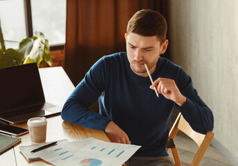 Poster - Thoughtful Millennial Guy Looking Through Data Graphs And Charts Thinking On Business Startup Sitting In Coworking Workspace. Selective Focus