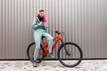 young hipster girl with dreadlocks in warm clothes uses a smartphone on a bicycle against the background of a gray wall, student cyclist typing online in cold weather in autumn