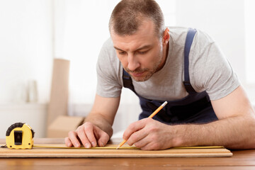 Fototapete - Carpenter At Work. Mature Craftsman Working Making Furniture Measuring Wooden Board Indoors, Wearing Blue Coverall Uniform. Carpentry Profession And Service. Selective Focus