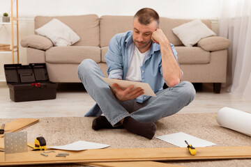 Puzzled Middle-Aged Man Reading Furniture Assembly Manual In Confusion Installing Shelf Sitting Among Self-Assembly Kit Tools And Wooden Parts On Floor At Home. Furniture Assembling Problem