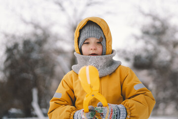 Young child wearing a bright yellow winter playing with a snowball maker in a snowy landscape during a cold winter day. Boy makes snowballs using a yellow plastic Snowball Maker Tool