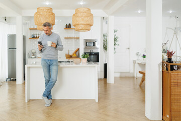 Mature man standing at a kitchen counter, holding a smartphone and coffee mug, looking focused while reading or browsing at home.