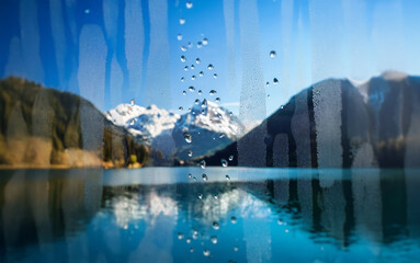 Poster - A view of a snow capped mountain range reflected in a still blue lake, viewed through a window covered in water droplets.