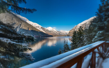 Poster - A serene mountain lake view framed by snow-covered evergreens and a wooden railing on a bright, sunny winter day.