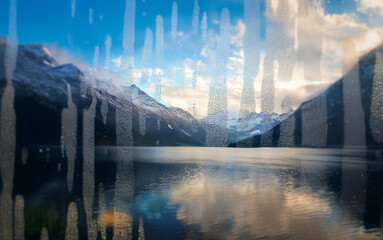 Poster - A scenic mountain lake view through a rain-streaked window, with the sun setting behind snow-capped peaks.